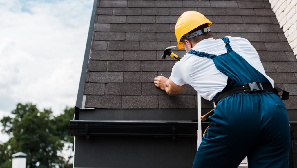 A roofer stands on a ladder with a hammer and nail in hand, repairing a shingle roof.