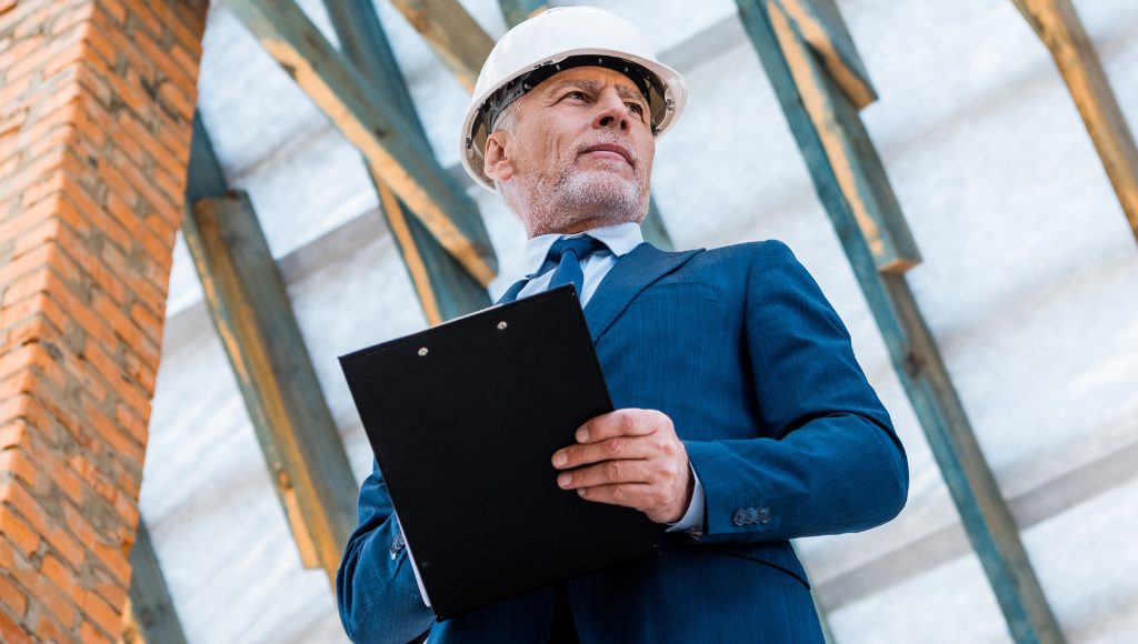 A professional in a blue suit and white hard hat holds a clipboard and inspects a roof.