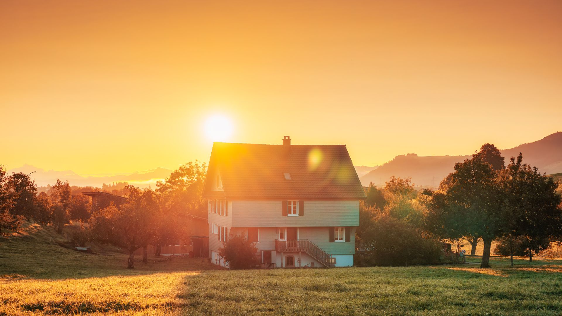 The sun rises over a rural house surrounded by trees. The UV rays shine directly onto the asphalt shingles.