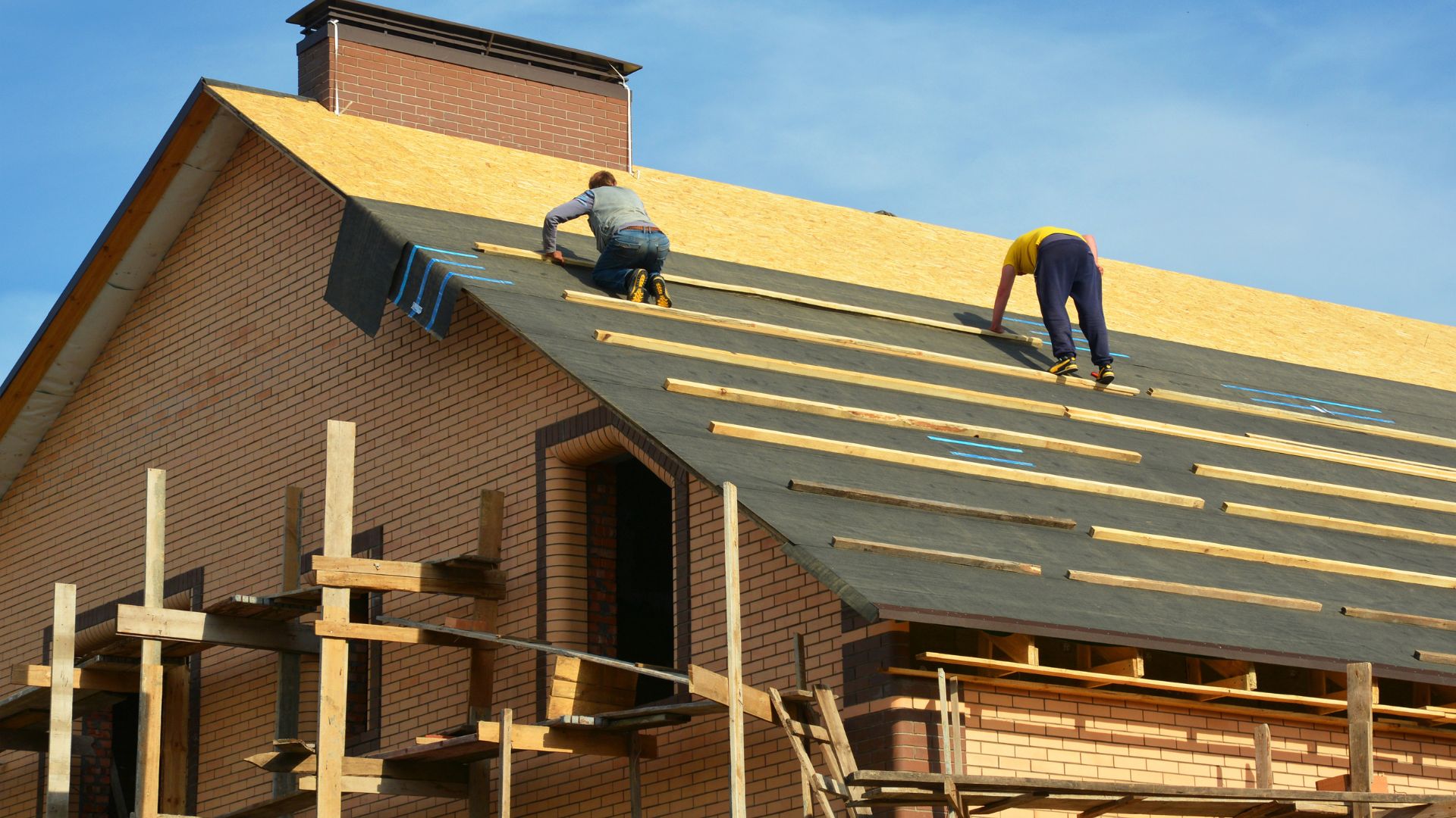 Roofing contractors are installing underlayment and a waterproofing membrane on the roof deck and the plywood sheathing of a large brick house under construction, with scaffolding.